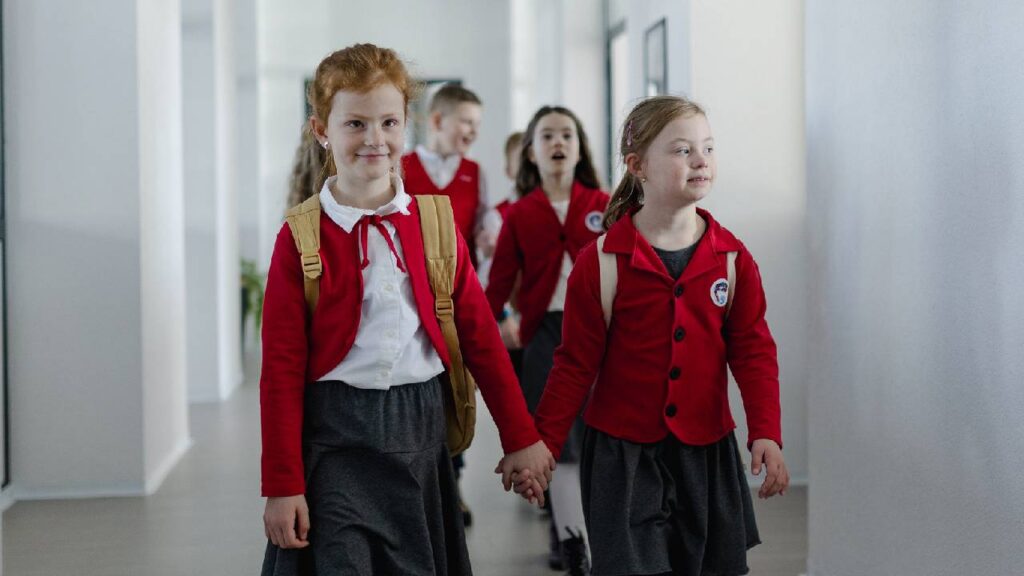 Happy schoolgrirl with Down syndrome in uniform holding hands with her classmate walking in scool corridor with classmates behind them.